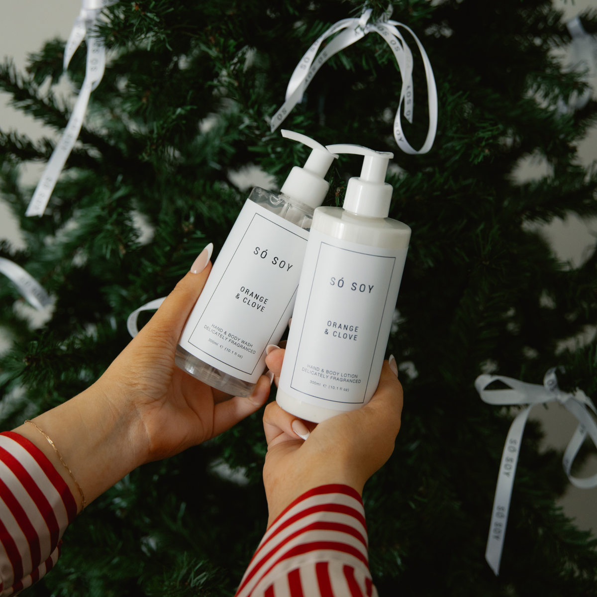 Two pump bottles of SO SOY Orange & Clove hand wash and hand lotion held by hands wearing a red and white striped shirt, set against a Christmas tree background with white ribbon ornaments.