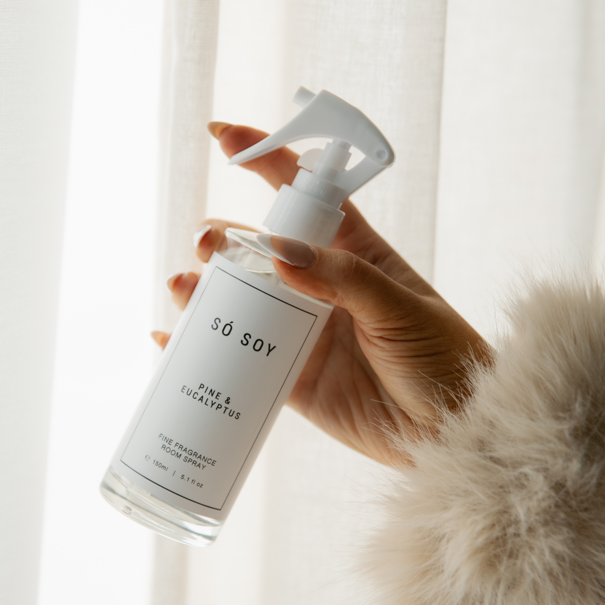 A hand holding a clear spray bottle of So Soy Pine & Eucalyptus fine fragrance oil spray against a soft white curtain background.