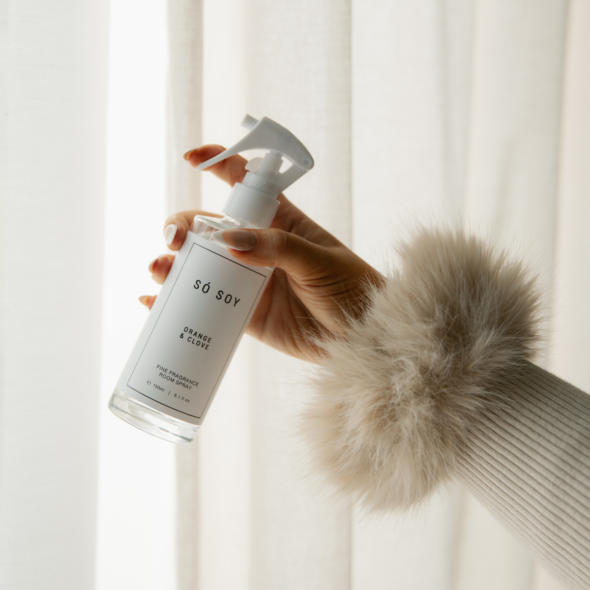 A hand wearing a beige faux-fur cuffed sweater holds a clear glass spray bottle of SÓ SOY Orange & Clove room spray against a soft white curtain background.