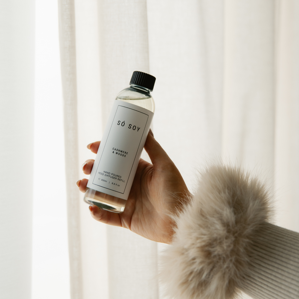 A hand wearing a fluffy beige sleeve holds a clear bottle of Só Soy cashmere & woods diffuser refill against a soft white curtain background.