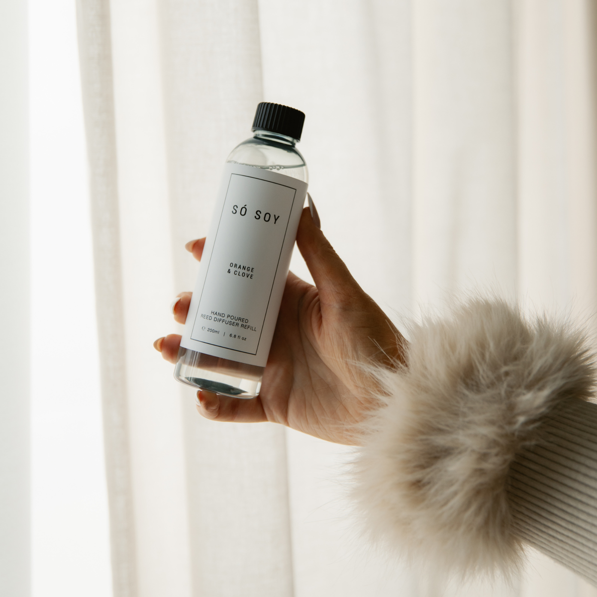 A hand holding a clear bottle of Só Soy Orange & clove reed diffuser refill against a soft, bright background of white curtains. The bottle has a white label with black text and a black cap, and the person holding it is wearing a beige faux-fur trimmed sweater.