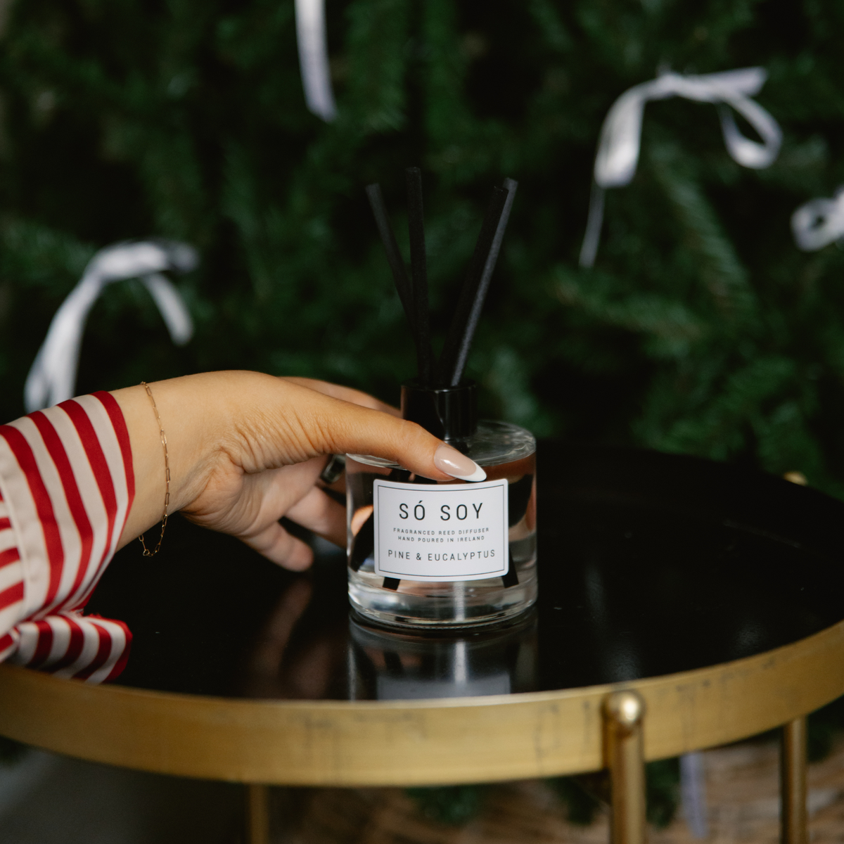 A hand wearing a red and white striped sleeve holds a glass reed diffuser bottle labeled 'SÓ SOY' on a black round table with a gold frame, with a blurred green holiday tree in the background.