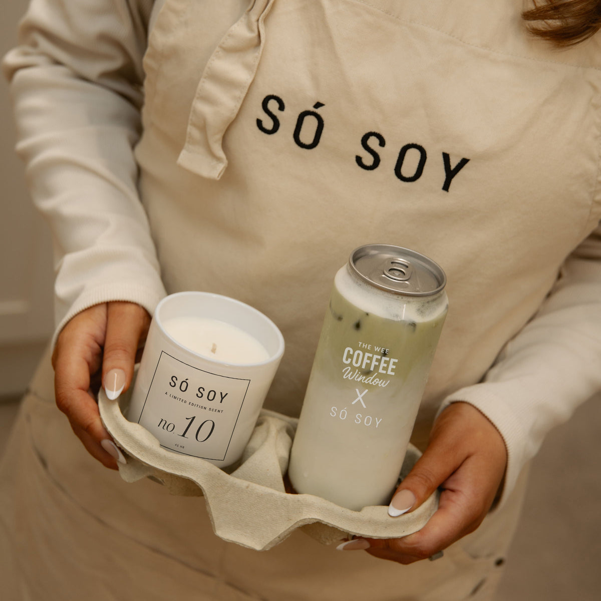 A person wearing a beige apron with 'SÓ SOY' printed in black holds a soy candle and a coffee-flavored can from SÓ SOY in a warm indoor setting.