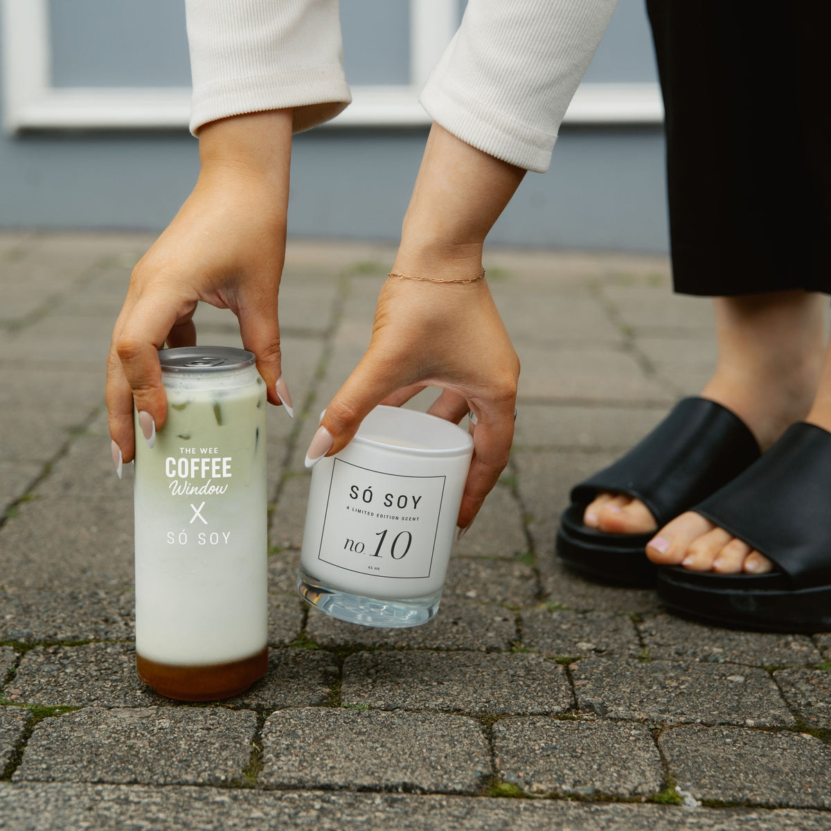 A person outdoors on a paved surface holds two glass containers: a tall frosted can labeled 'COFFEE x SO SOY' with a white top, and a small clear jar labeled 'SO SOY no. 10' with a white lid.
