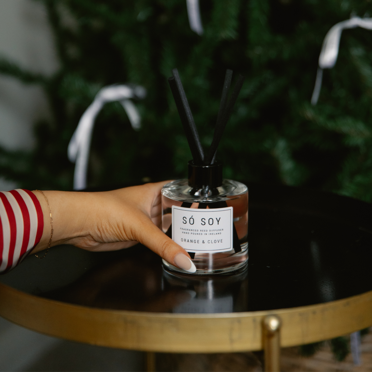 A hand in a red and white striped sleeve holds a reed diffuser bottle labeled 'SÓ SOY' on a black round table with a gold frame, with a blurred green holiday tree in the background.