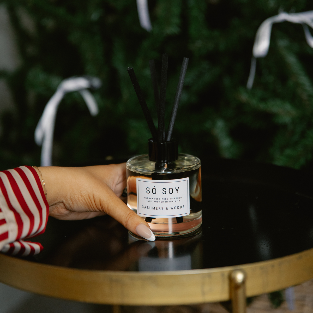 A hand in a red and white striped sleeve holds a glass reed diffuser bottle labeled 'SÓ SOY' on a black table with a gold frame, with a blurred green holiday tree in the background.
