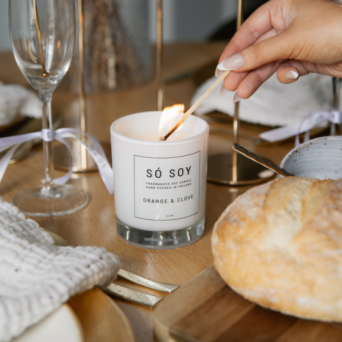 A hand lighting a candle with a wooden match. The candle is in a clear glass jar with white wax and a black label that reads 'SÓ SOY' and 'ORANGE & CLOVE'.