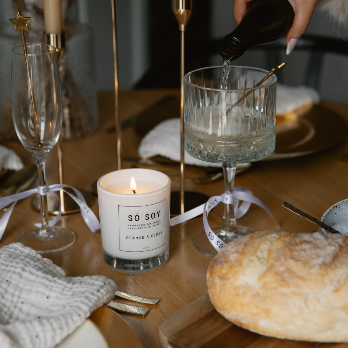 A styled dining table scene with a lit white candle labeled “SÓ SOY” in the center, a loaf of bread in the foreground, and a hand pouring liquid from a dark bottle into a glass. Gold-toned tableware, ribbons, and candles create a warm, lifestyle-oriented setting.