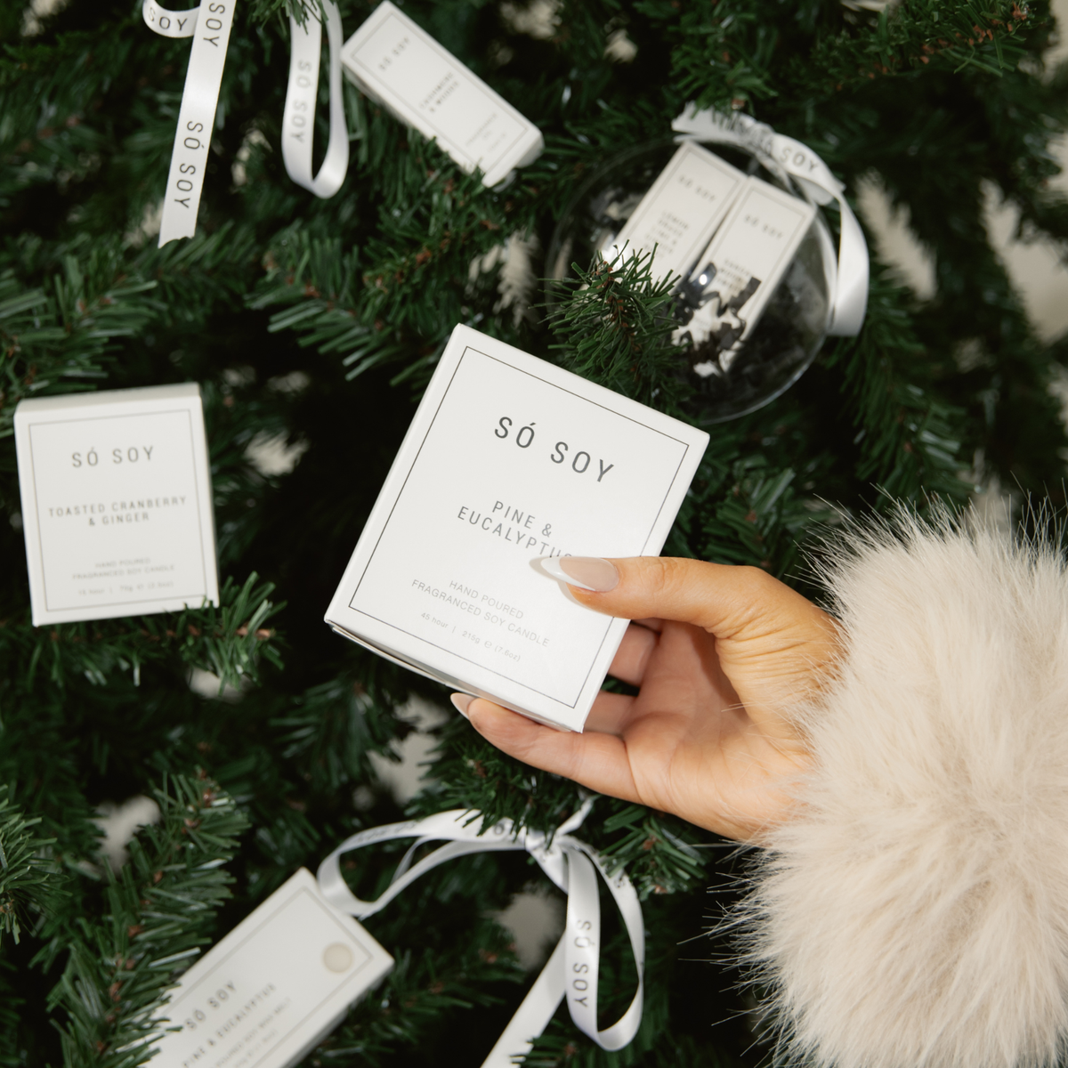 A hand wearing a fluffy cream sleeve holds a square white box of SÓ SOY Pine & Eucalyptus scented candles, surrounded by a decorated Christmas tree with other SÓ SOY ornaments and tags.