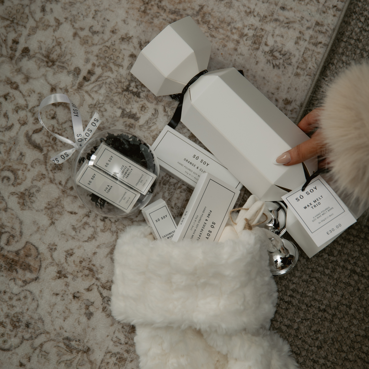 A hand holds a white cylindrical gift box tied with a black ribbon on a patterned rug, alongside white packaging cards, a clear ornament filled with black items, and a fluffy white stocking.