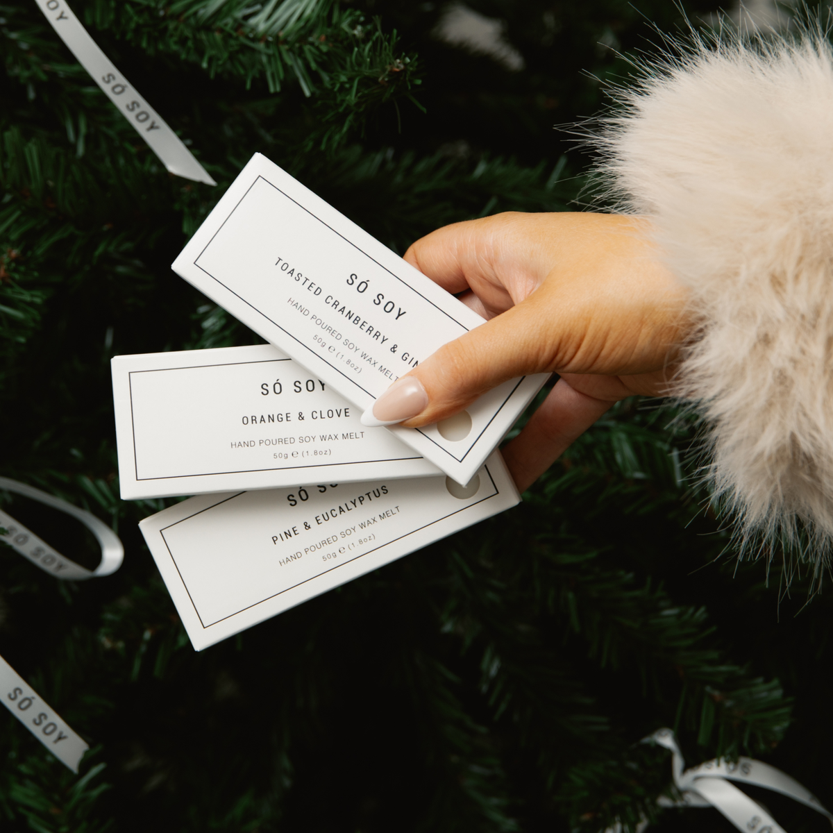 A hand with manicured nails holds three rectangular wax melts from the brand Sø Soy against a dark green Christmas tree background with white ribbons. The wax melts are in different scents: Toasted Cranberry & Ginger, Orange & Clove, and Pine & Eucalyptus.
