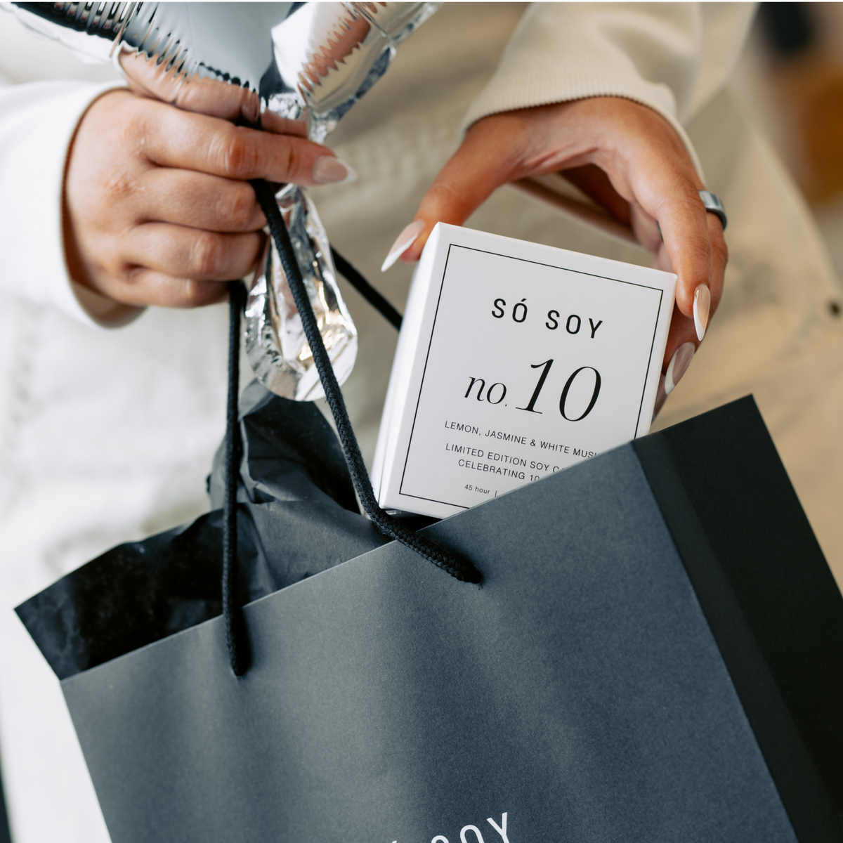 A person holds a small white box labeled 'só soy no 10' above a dark gray shopping bag with black handles. The person wears a light beige coat, and silver foil balloons are visible in the background.