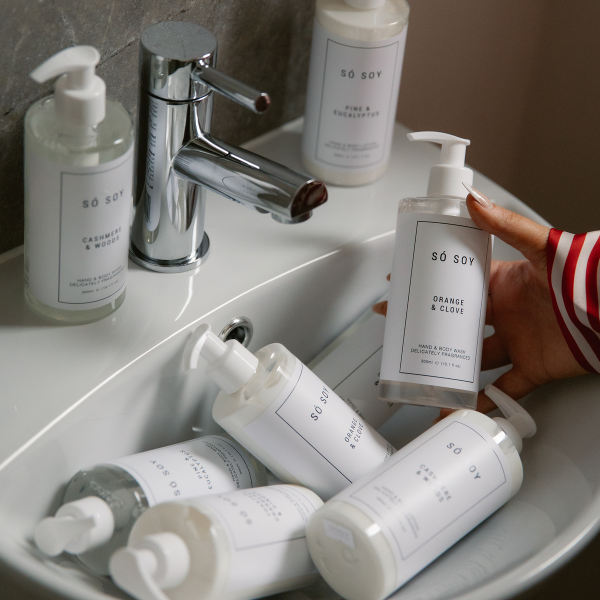 A hand in a red and white striped sleeve holds a bottle of SÓ SOY Orange & Clove hand wash next to a chrome faucet in a white sink, with several other SÓ SOY bottles arranged around it.
