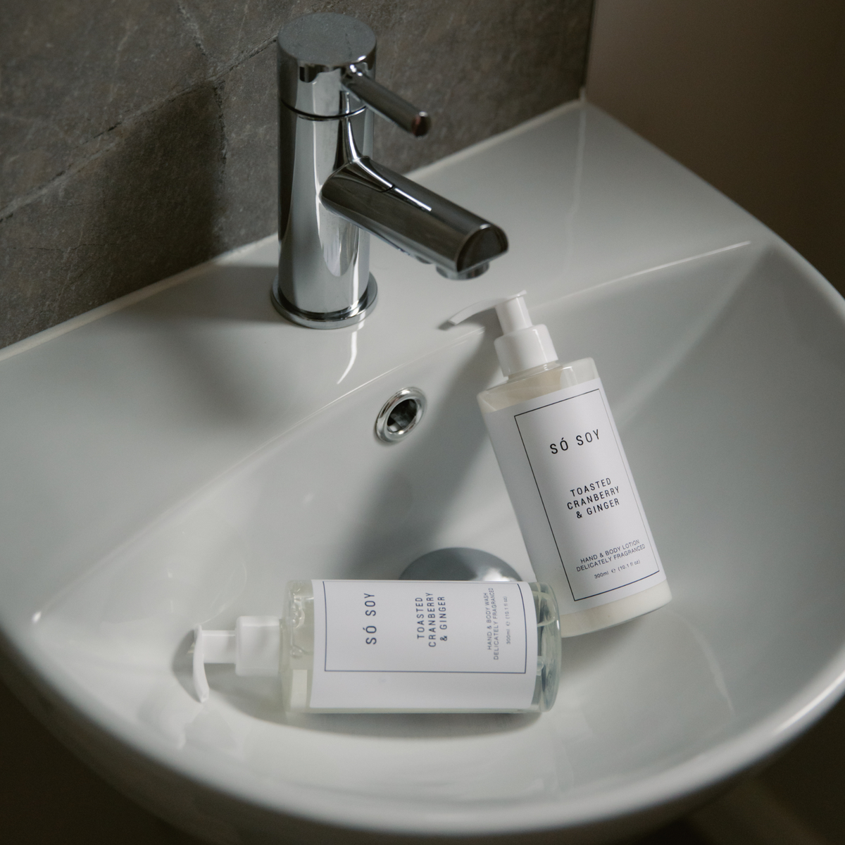 Two white pump bottles from the brand so soy, one hand wash and one body wash, displayed on a white ceramic bathroom sink next to a chrome faucet.