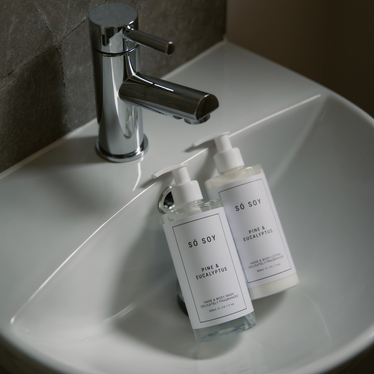 Two pump bottles of SÓ SOY hand and body wash resting in a white ceramic bathroom sink next to a chrome faucet.