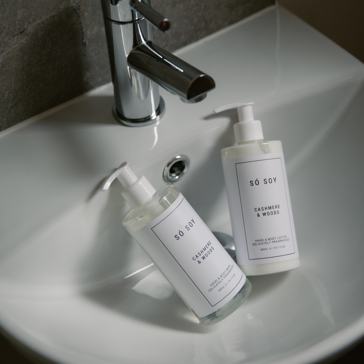 Two white pump bottles labeled SÓ SOY Cashmere & Woods body wash and hand lotion resting on a white ceramic bathroom sink with a chrome faucet.
