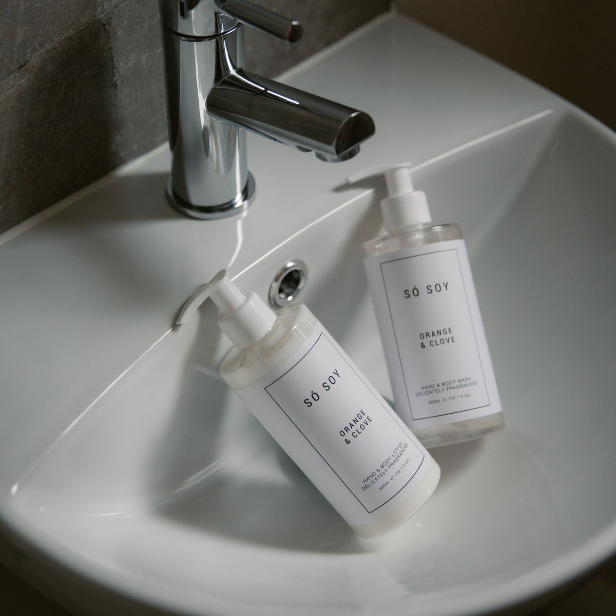 Two clear pump bottles of S°SOY hand and body wash with white labels, placed on a white ceramic bathroom sink next to a chrome faucet.