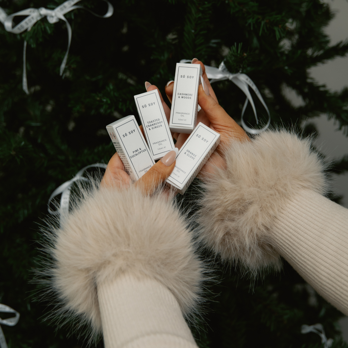 Four small white rectangular Só Soy fragrance oils for aroma diffusers held in two hands wearing fluffy cream faux-fur wrist cuffs and ribbed cream sleeves, with a dark green Christmas tree and white ribbon in the background.