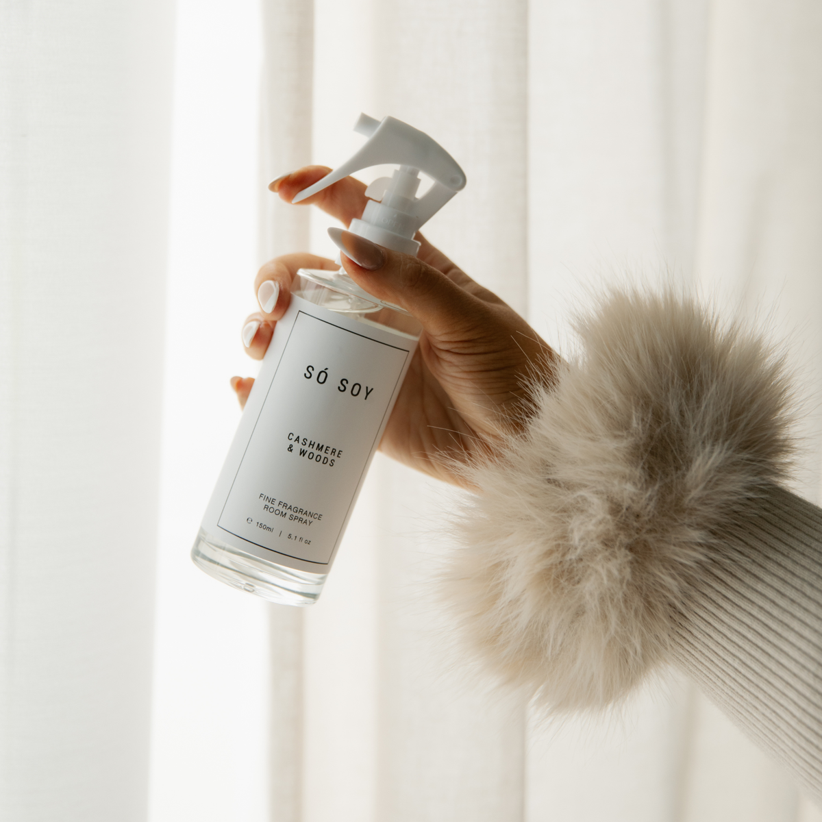 A hand holding a clear spray bottle of Só Soy Cashmere & Woods Fine Fragrance room spray, set against a soft, bright background of white curtains. The hand is adorned with a fluffy beige faux-fur sleeve cuff.
