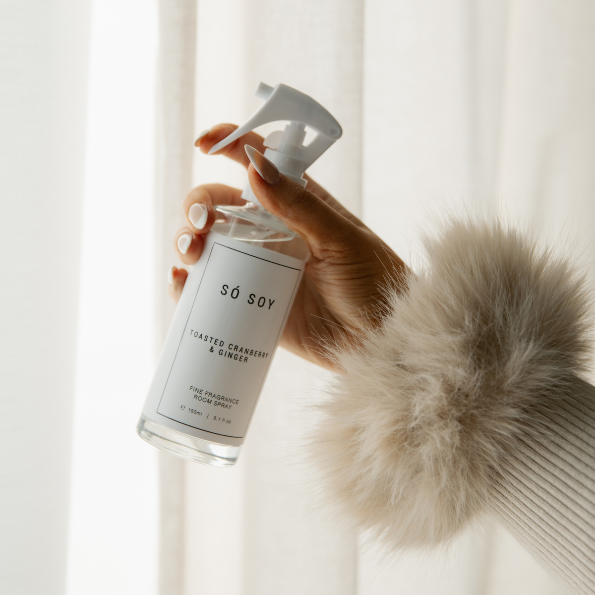 A hand holding a clear spray bottle of Sö Soy Fine Fragrance room spray with a white label and white spray top, set against a soft white curtain background with a beige faux-fur sleeve visible.