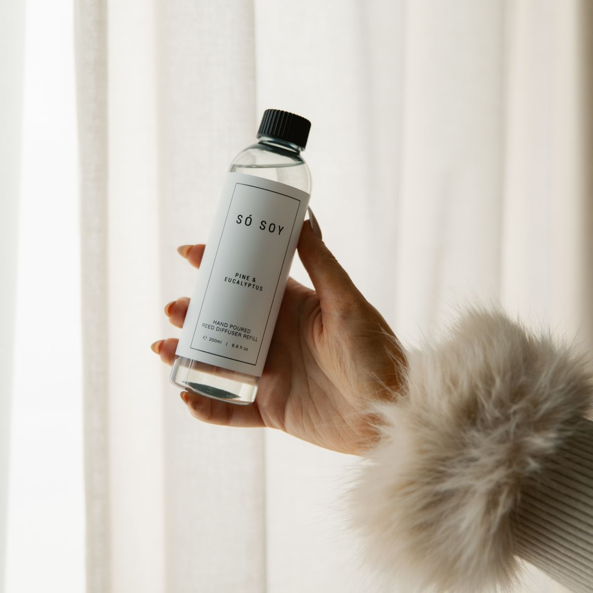 A hand holding a clear bottle of SO SOY Pine & Eucalyptus reed diffuser refill against a soft, bright background with white curtains and a fluffy cream sleeve visible.