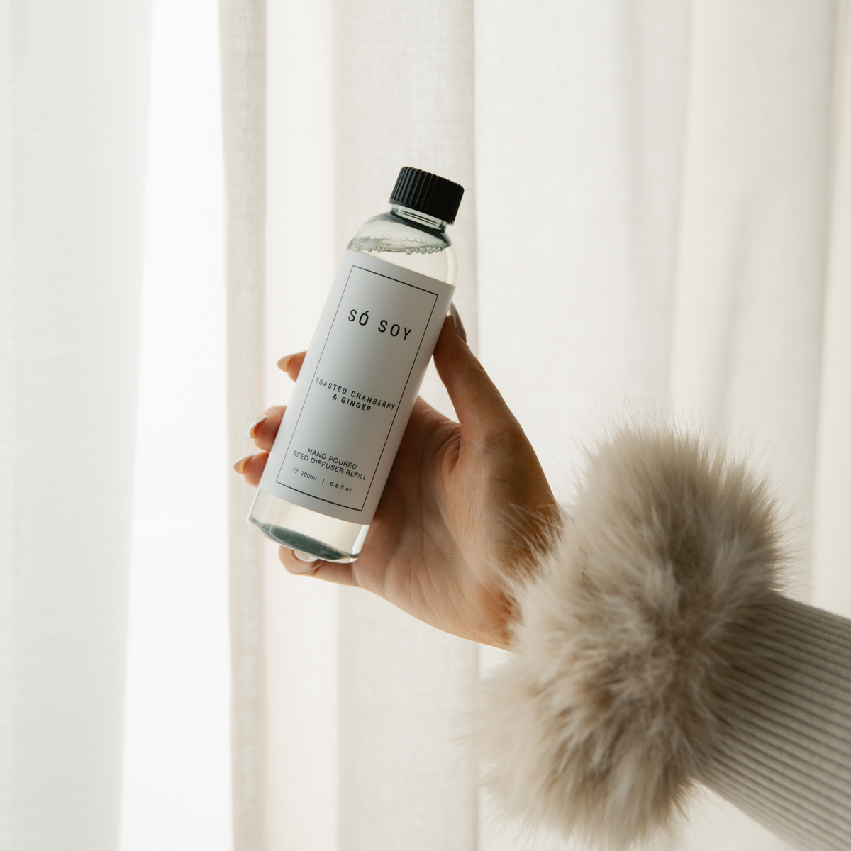 A hand wearing a fluffy cream faux-fur sleeve holds a clear bottle of SÓ SOY diffuser refills against a soft white curtain background.