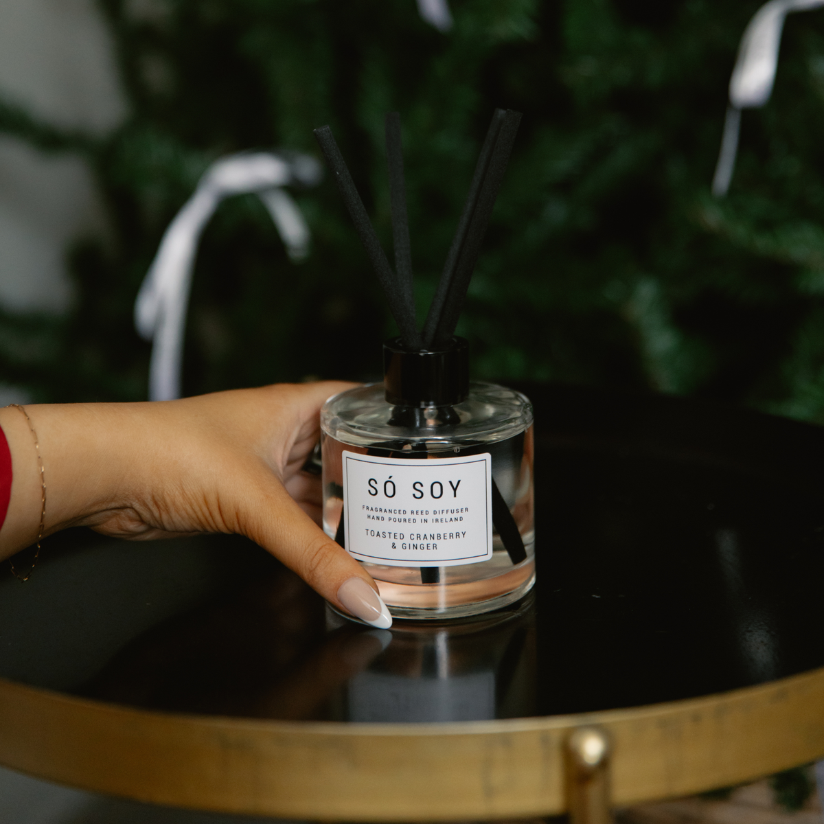 A hand holding a clear glass reed diffuser bottle labeled 'SÓ SOY' with black reeds and a black cap, set on a glossy black table with a blurred green holiday tree in the background.