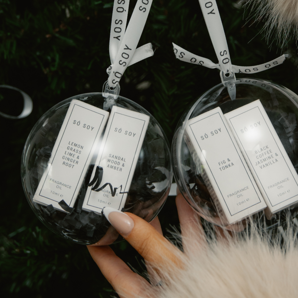 Two clear round Christmas ornaments hanging on a dark green Christmas tree, each containing two small white fragrance oils bottles with silver ribbons printed with 'SO SOY'. Hands in cream faux fur sleeves are holding the ornaments.
