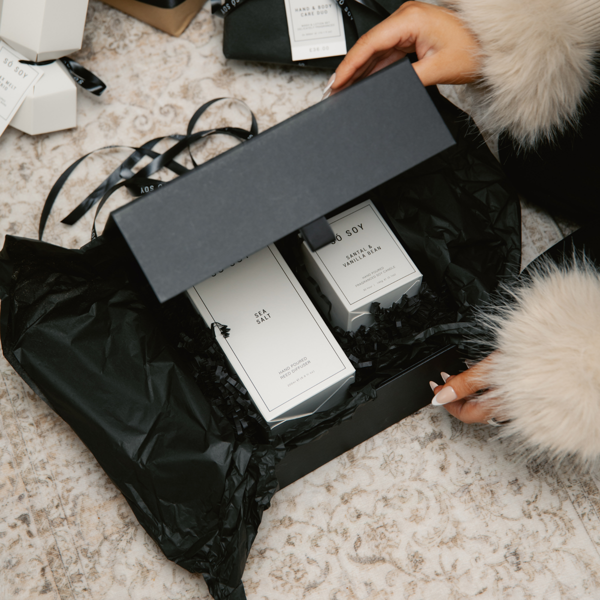 A person's hands, clad in a white faux fur sleeve, open a black gift box containing two white home fragrance products on a patterned beige rug.