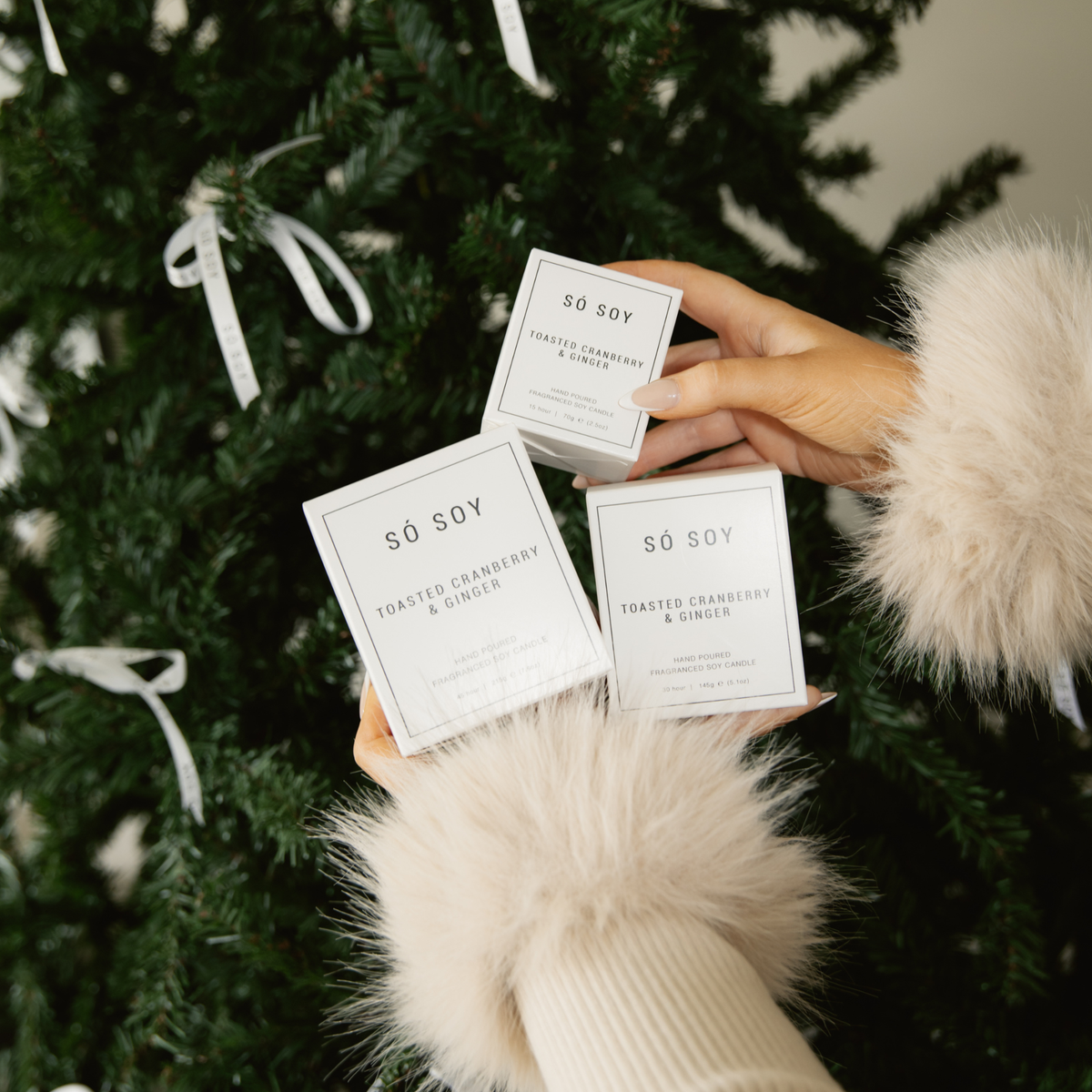 Three small white square boxes of So Soy candles, including Toasted Cranberry & Ginger, held by hands wearing fluffy cream faux fur sleeves in front of a decorated Christmas tree.