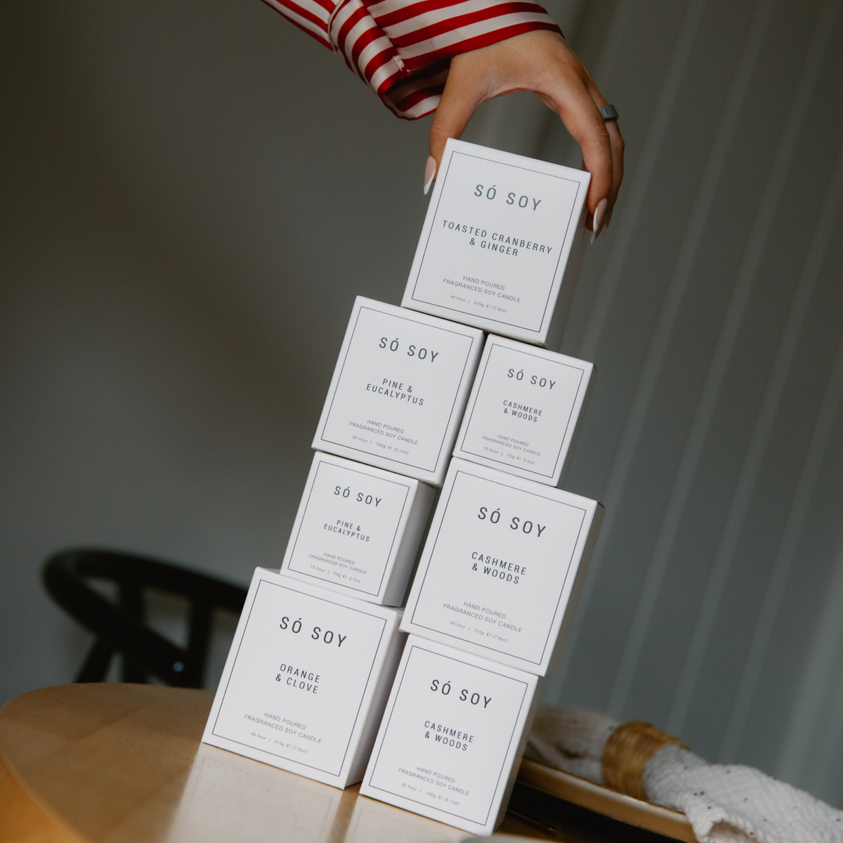 A hand with manicured nails stacks multiple white rectangular boxes of Sø Soy candles on a wooden table. .