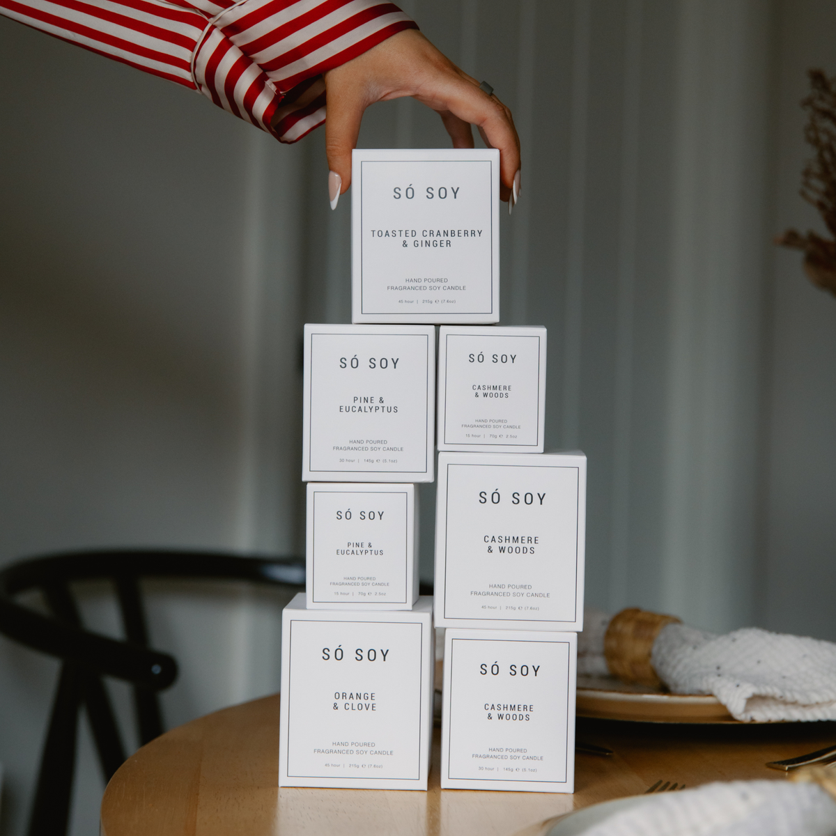 A hand wearing a red and white striped sleeve stacks several white rectangular boxes of SÓ Soy festive candles