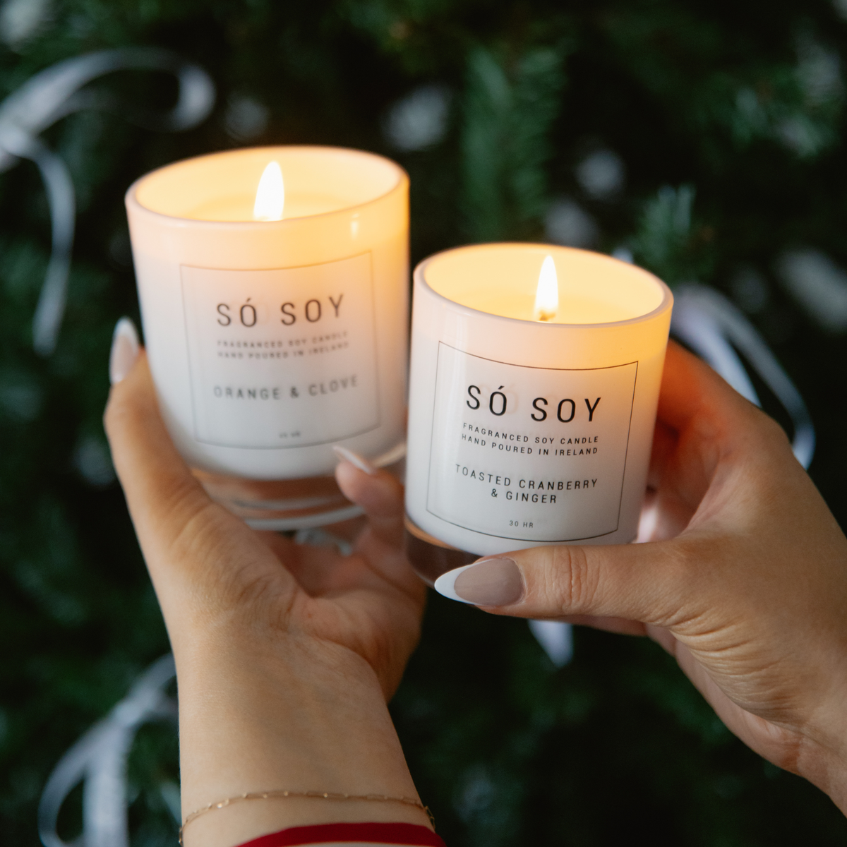 Two hands holding lit SÓ SOY soy candles in glass jars, with one candle labeled Orange & Clove and the other labeled Toasted Cranberry & Ginger, against a dark green holiday background.