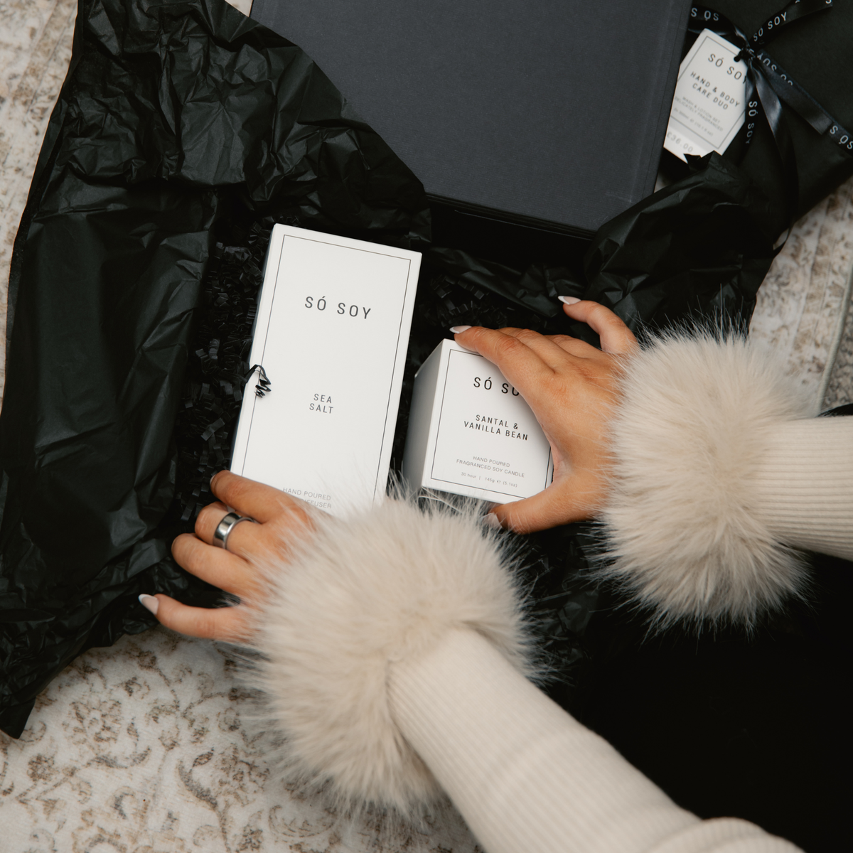 A close-up of a gift box opened with black tissue paper, containing two white product boxes labeled SÖ SOY. A person's hands with cream faux-fur trimmed sleeves are visible holding the boxes.