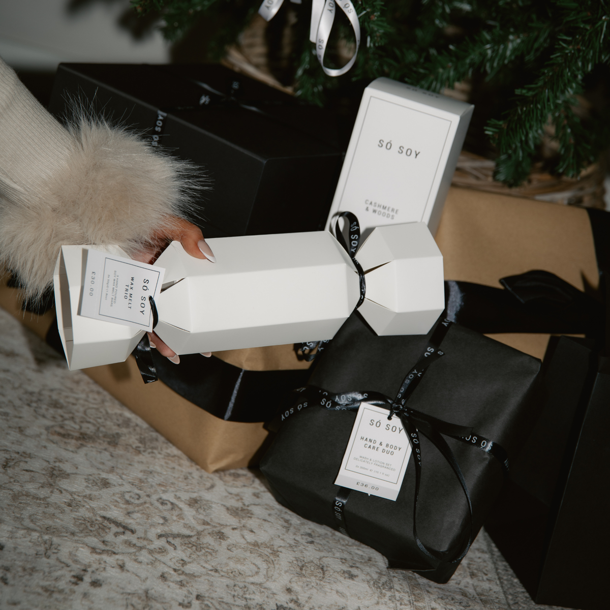 A hand wearing a cream faux-fur sleeve holds a So Soy Wax Melt trio cracker tied with a black ribbon. Several other gift boxes and wrapped presents are arranged on a patterned rug beneath a Christmas tree.