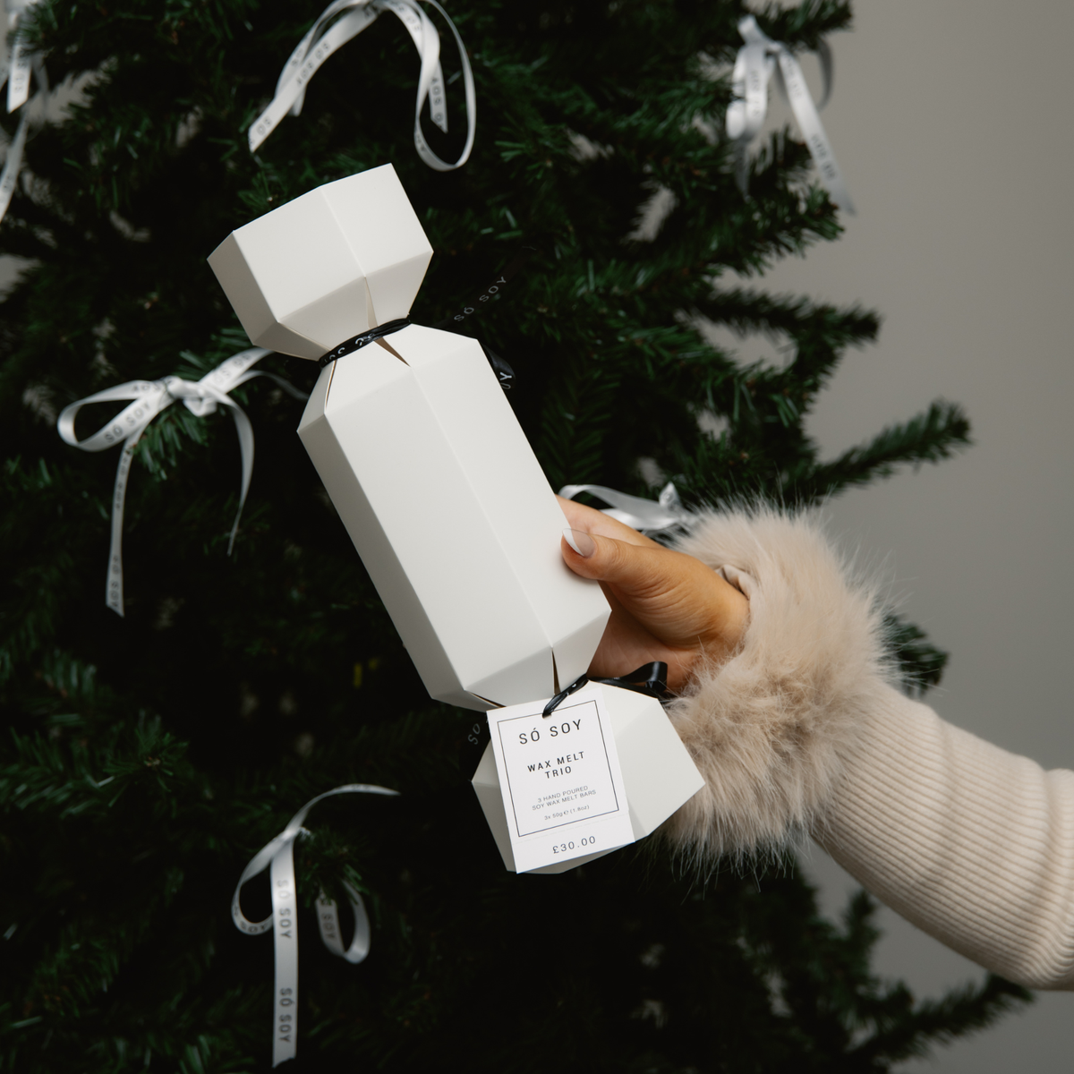 A hand wearing a cream faux-fur sleeve holds a white trio wax melt Christmas cracker, with a hanging tag that reads 'SO SOY'. The ornament is positioned in front of a dark green Christmas tree decorated with white ribbons.