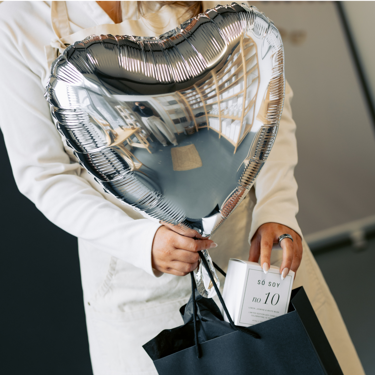 A person in a white shirt holds a large reflective silver heart-shaped foil balloon and a black shopping bag containing a white box labeled 'SO SOY no 10'.